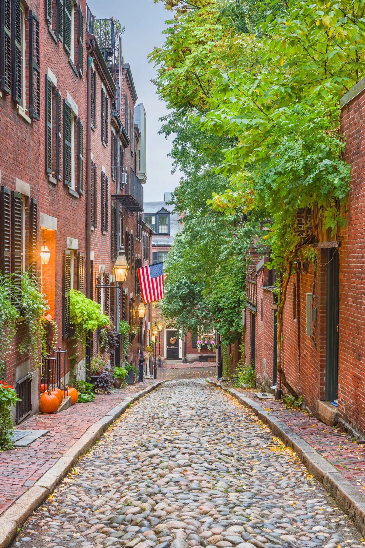 A cobblestone street in Boston’s Beacon Hill lined with red brick buildings, black shutters, and gas lanterns. Pumpkins and potted plants decorate the steps, and an American flag hangs from one of the homes.