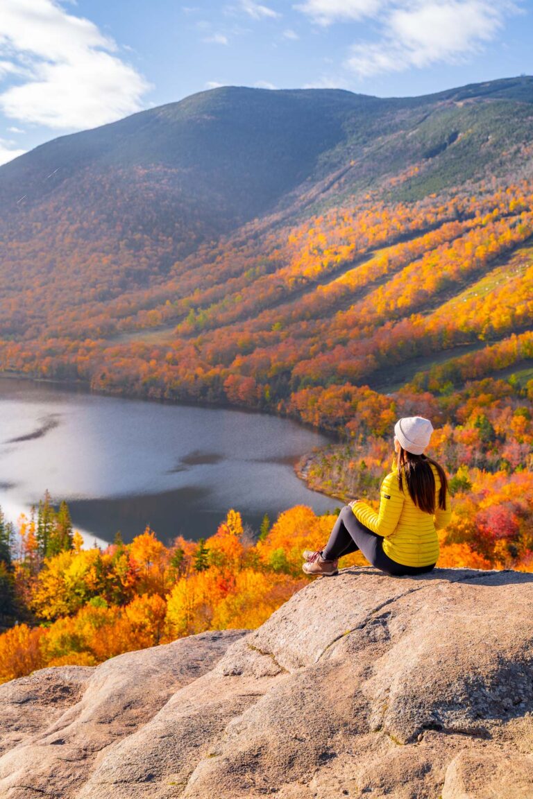 A woman in a yellow jacket and beanie sits on a rocky ledge at Artists Bluff in New Hampshire, overlooking a lake and hills covered in vibrant fall colors.