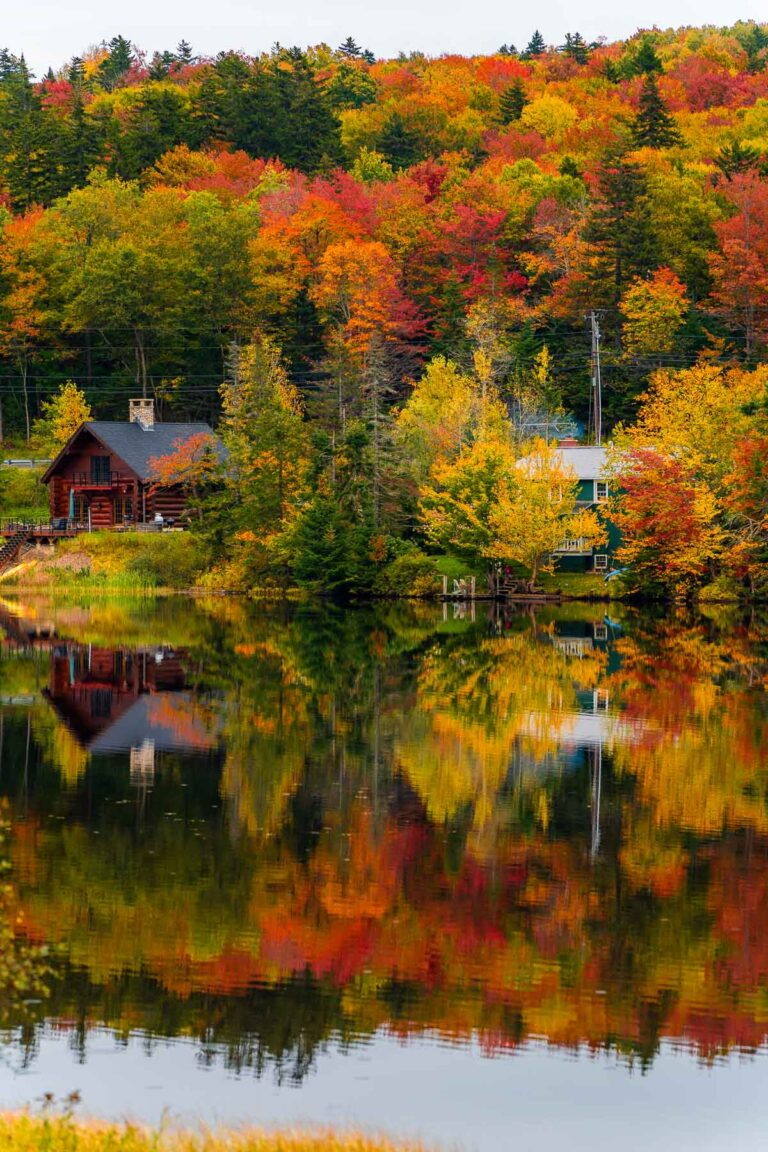Colorful fall foliage reflected in the still water of Big Pond in Vermont. A log cabin and another house sit near the shore surrounded by trees in shades of red, orange, and yellow.