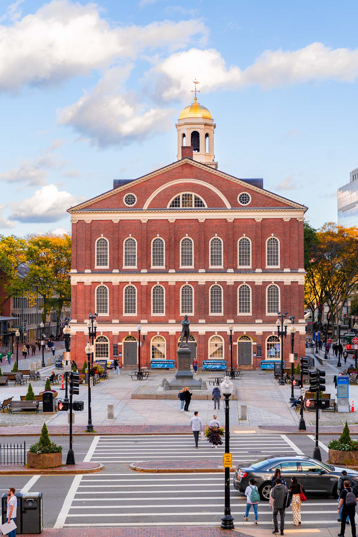 Looking straight at the front of Faneuil Hall in Boston, with people walking across the crosswalk and gathering around the statue in front.