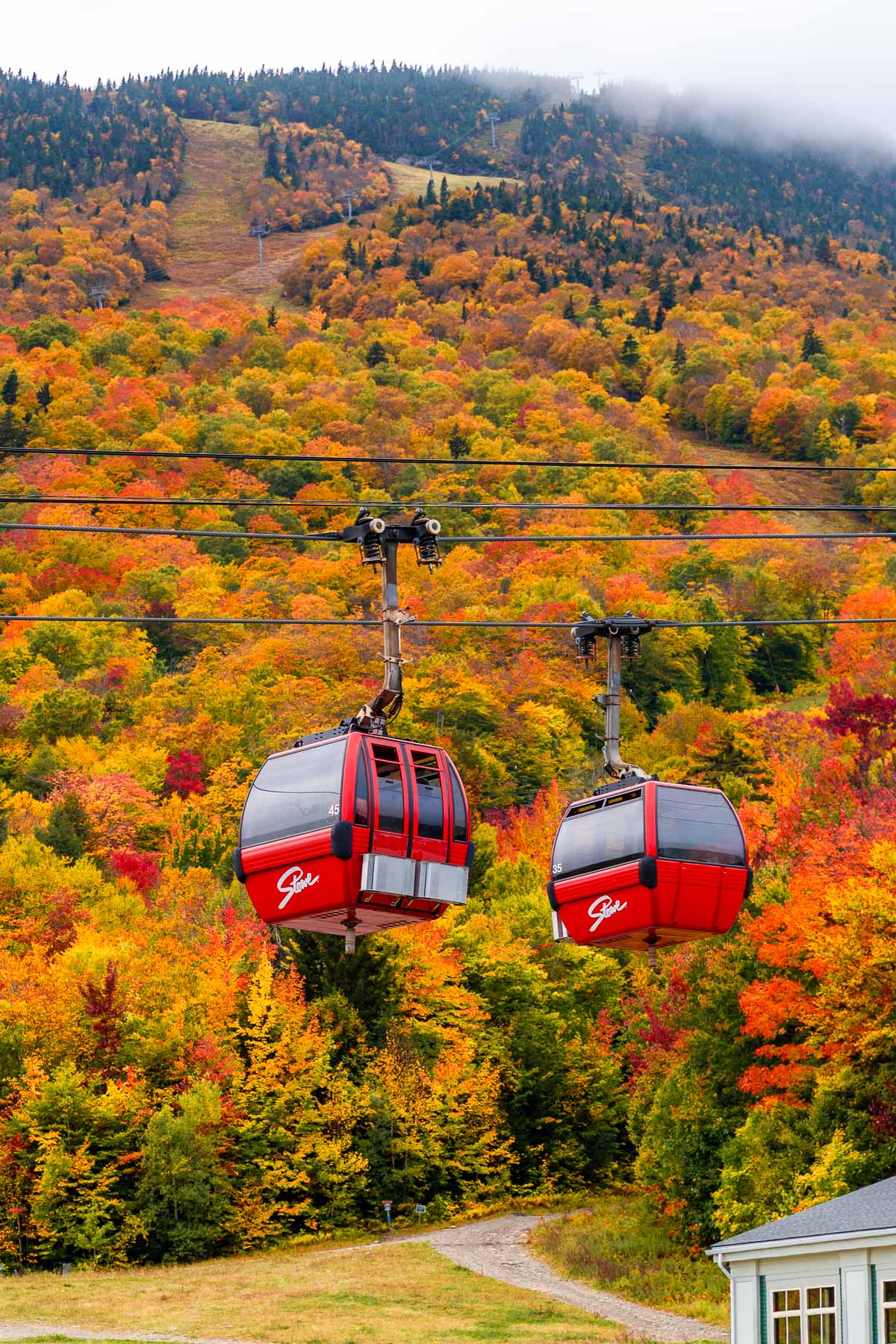 Two red gondolas on the Gondola SkyRide in Stowe, Vermont moving over a hillside covered in orange, yellow, and red fall foliage.