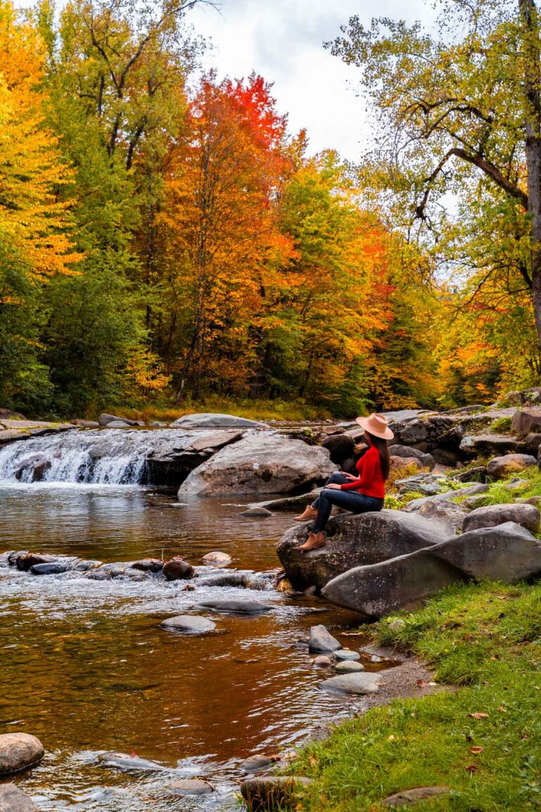 A woman in a red top and hat sitting on a rock by the river near Grist Mill Covered Bridge in Vermont, surrounded by colorful fall trees and a small waterfall.