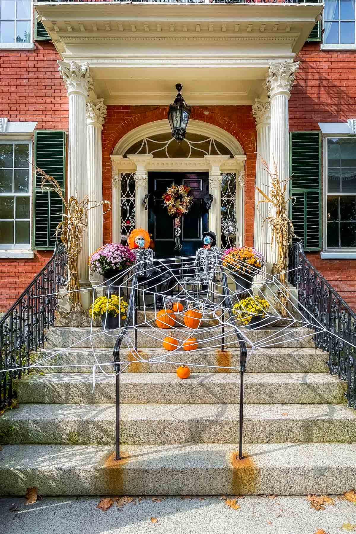 A brick house in Salem with a grand front entrance dressed up for Halloween. The steps are covered with a large white spiderweb and small pumpkins, while two skeletons in wigs and masks sit by the black front door surrounded by mums and corn stalks.