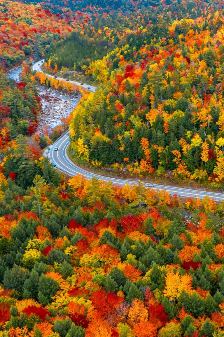 An aerial view of Kancamagus Highway in New Hampshire winding through hills covered in bright red, orange, and yellow fall foliage.