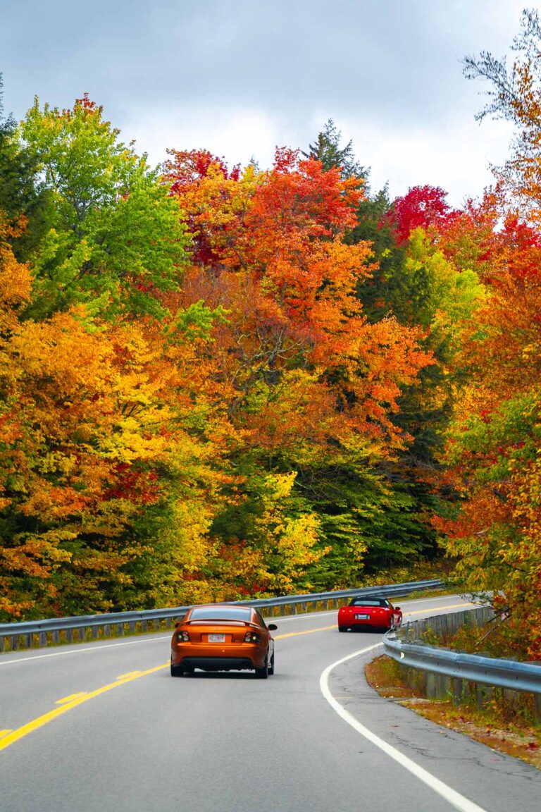 Two cars driving along Kancamagus Highway in New Hampshire with orange, yellow, and red autumn trees lining the road.