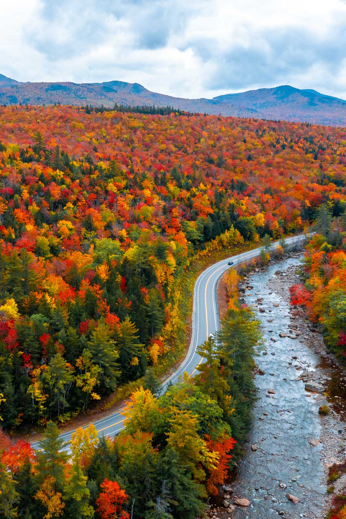 An aerial view of Kancamagus Highway in New Hampshire running alongside a rocky river with hills full of red, orange, and yellow fall trees.