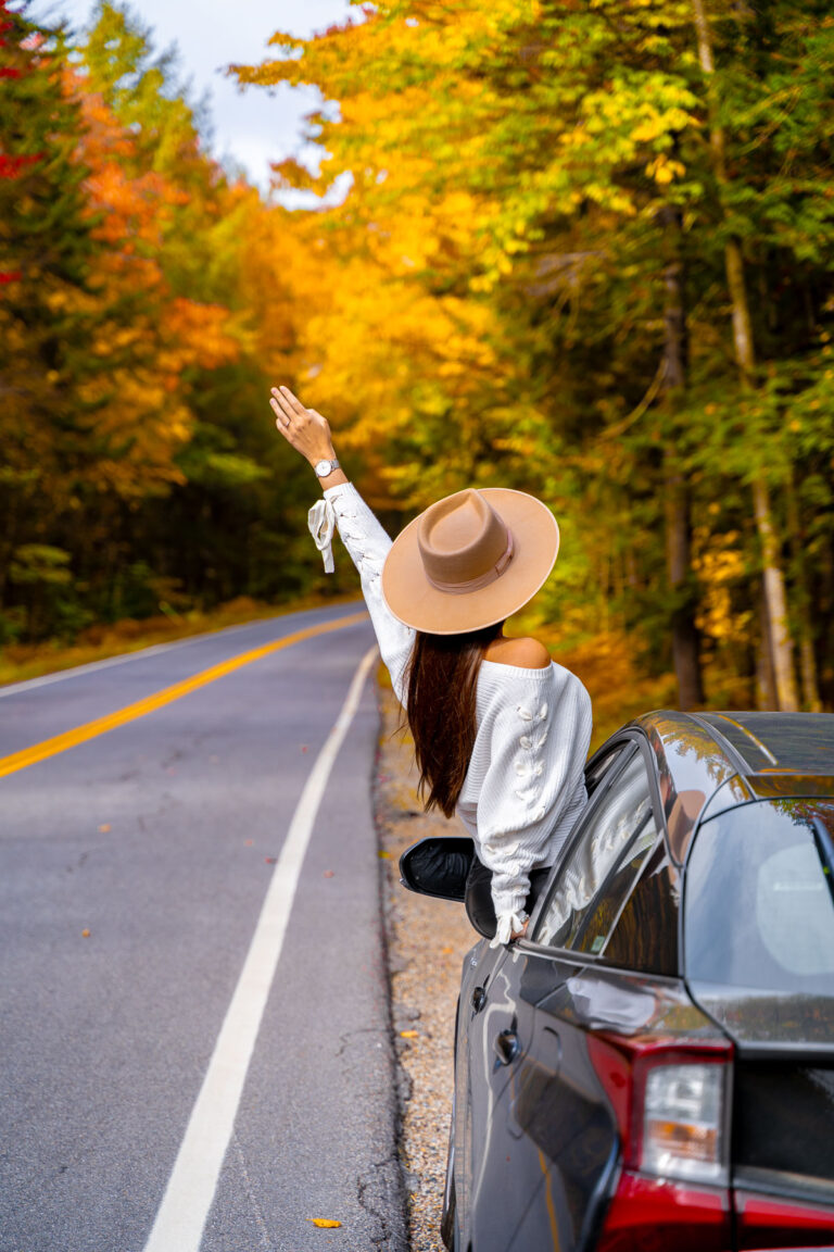 A woman in a tan hat and white sweater sitting on the window frame of a car parked along the Kancamagus Highway in New Hampshire. The road is lined with vibrant fall foliage in shades of yellow, orange, and red.