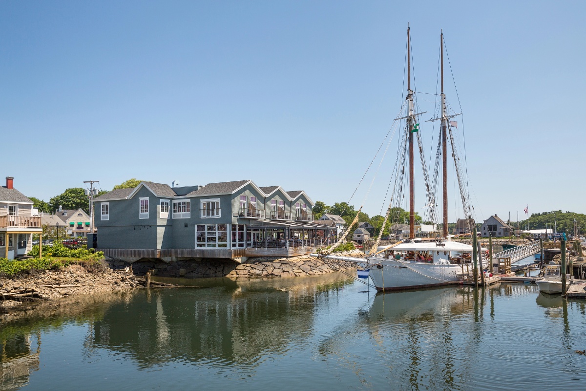A two-masted schooner docked beside a waterfront building in Kennebunkport, with calm waters and bright blue skies on a summer day.