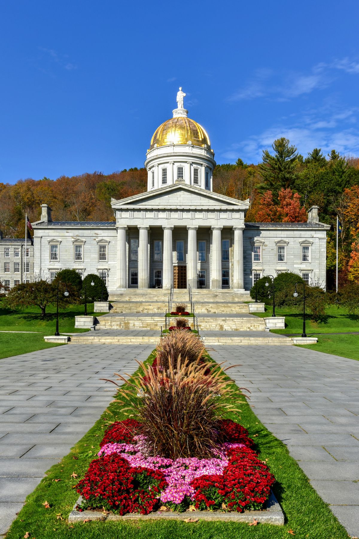 The Vermont State House in Montpelier with its golden dome, surrounded by fall foliage and a landscaped garden in front.