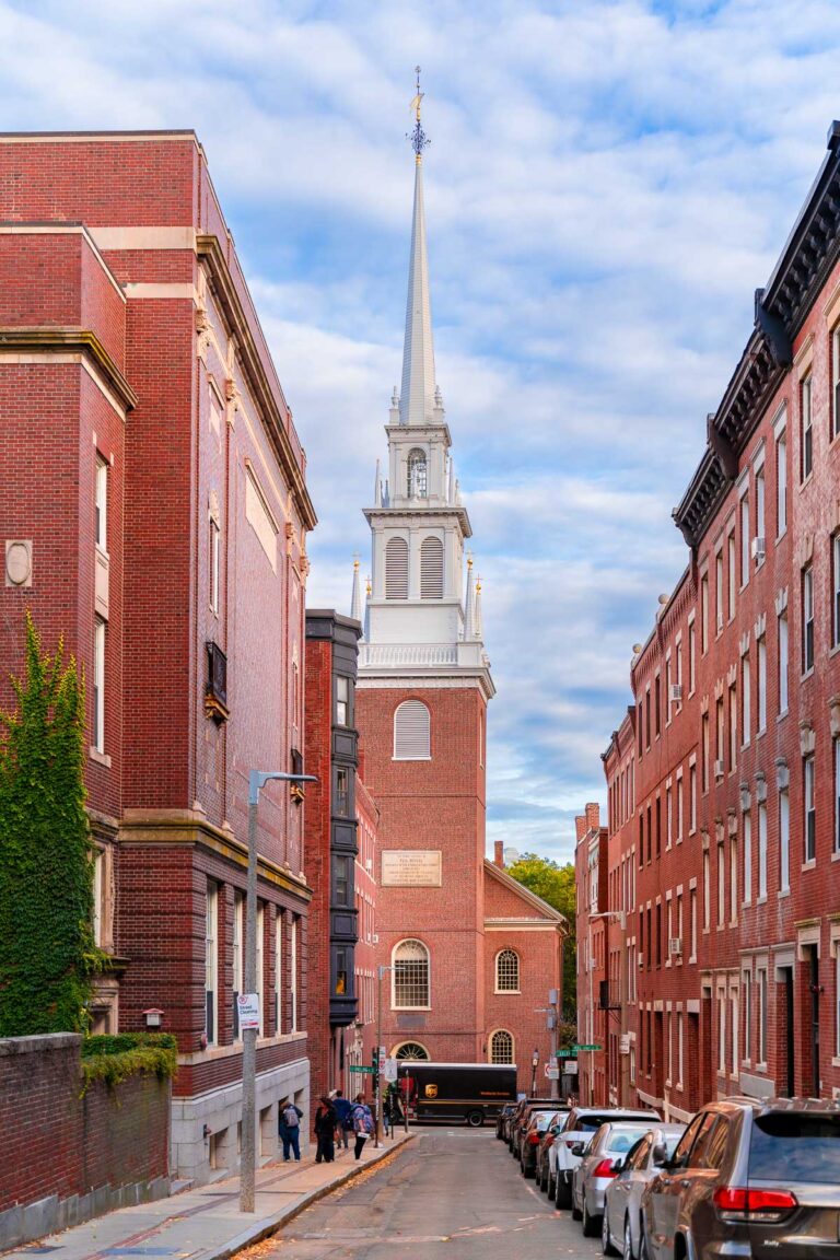 View of the Old North Church in Boston framed by red brick buildings and a narrow street, with a few people walking and cars parked along the curb.