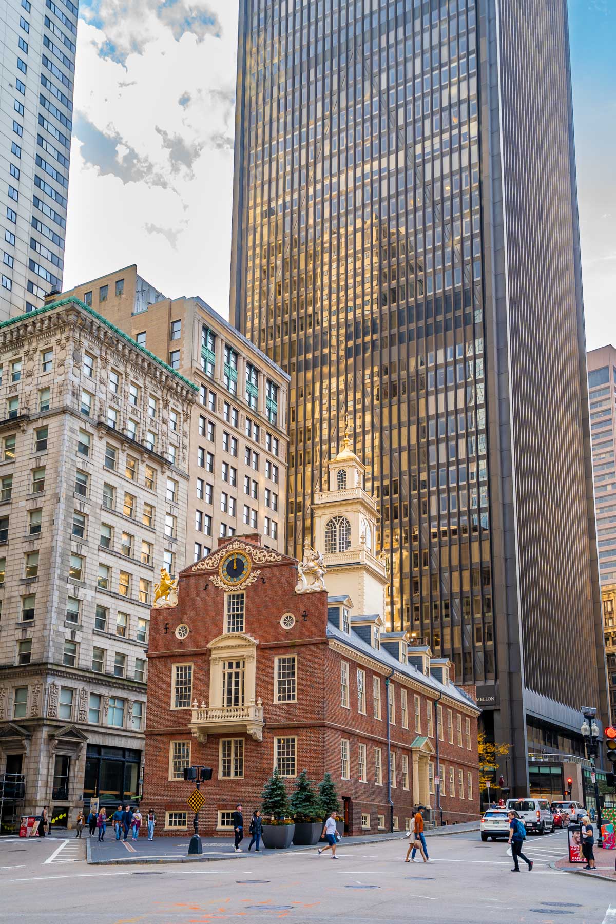 The Old State House in Boston stands out with its red brick facade and white trim, surrounded by towering modern skyscrapers and a busy intersection.