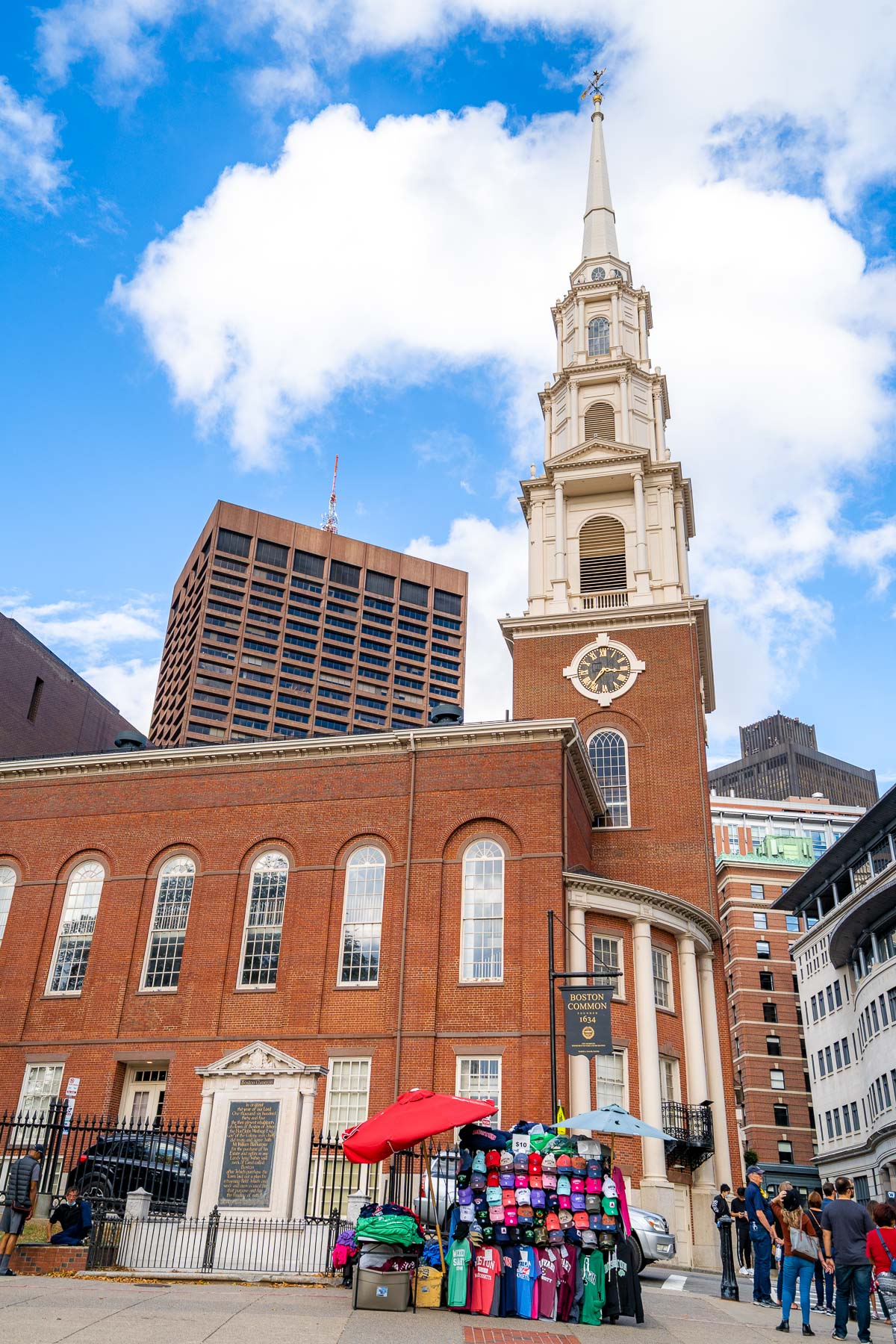 Park Street Church in Boston with its tall white steeple and red-brick facade, seen from the Boston Common entrance where a street vendor is set up in front.