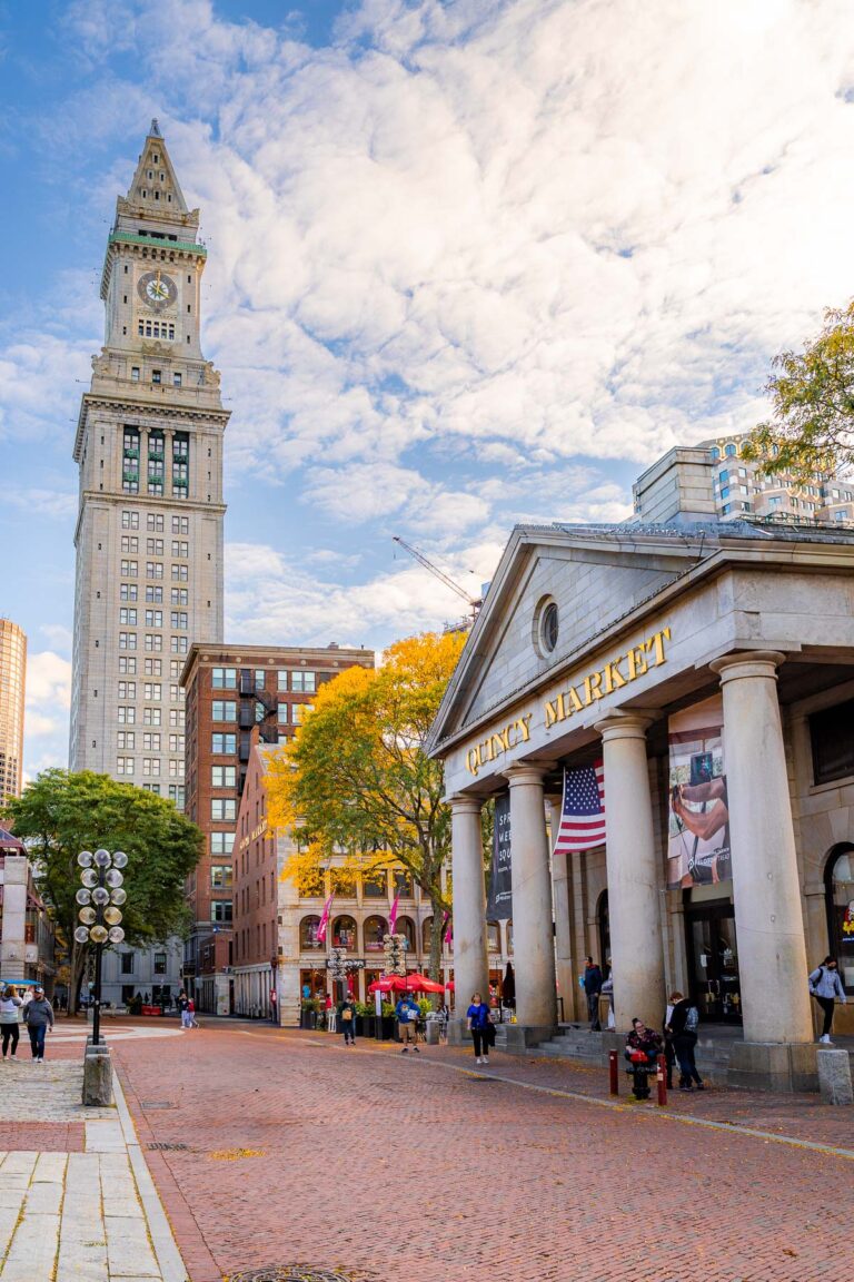 Quincy Market in Boston with its stone columns and American flag out front, framed by trees with fall leaves and the Custom House Tower in the background.