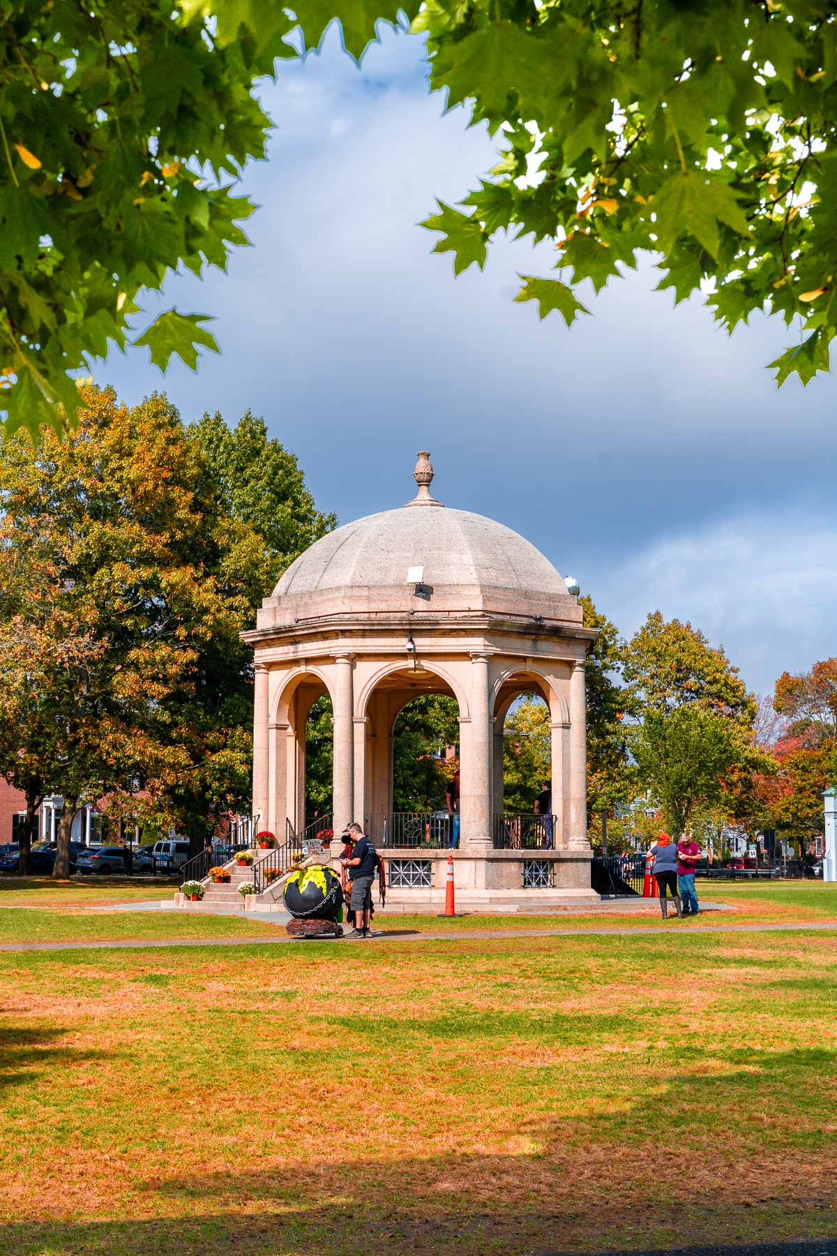 A sunny afternoon at Salem Common with people walking near the white gazebo, surrounded by green lawns and trees starting to turn with fall color.