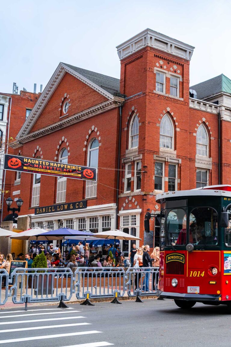 A red Salem Trolley passes in front of the old Daniel Low & Co. building on Essex Street, decked out with Halloween banners and packed with people enjoying the Haunted Happenings marketplace.