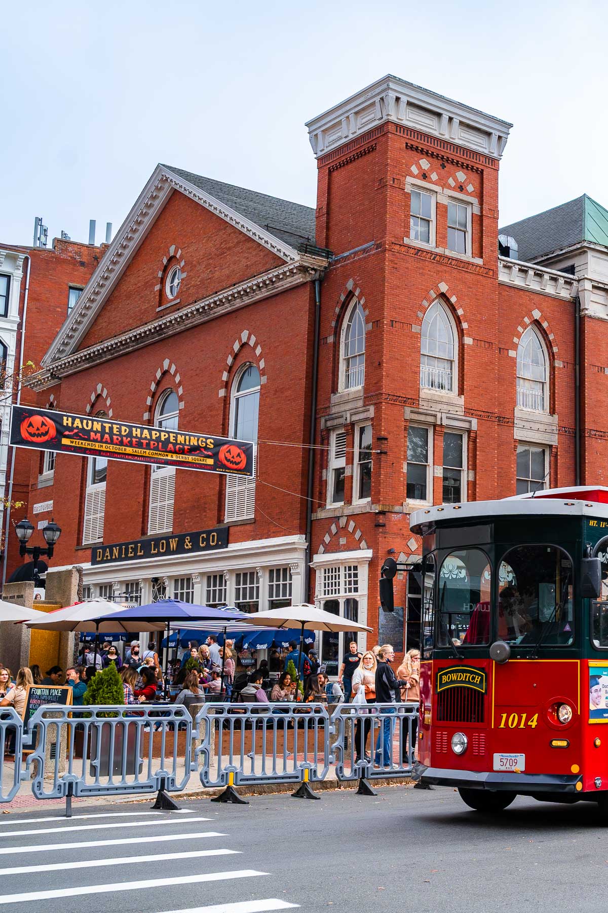 A red Salem Trolley passes in front of the old Daniel Low & Co. building on Essex Street, decked out with Halloween banners and packed with people enjoying the Haunted Happenings marketplace.