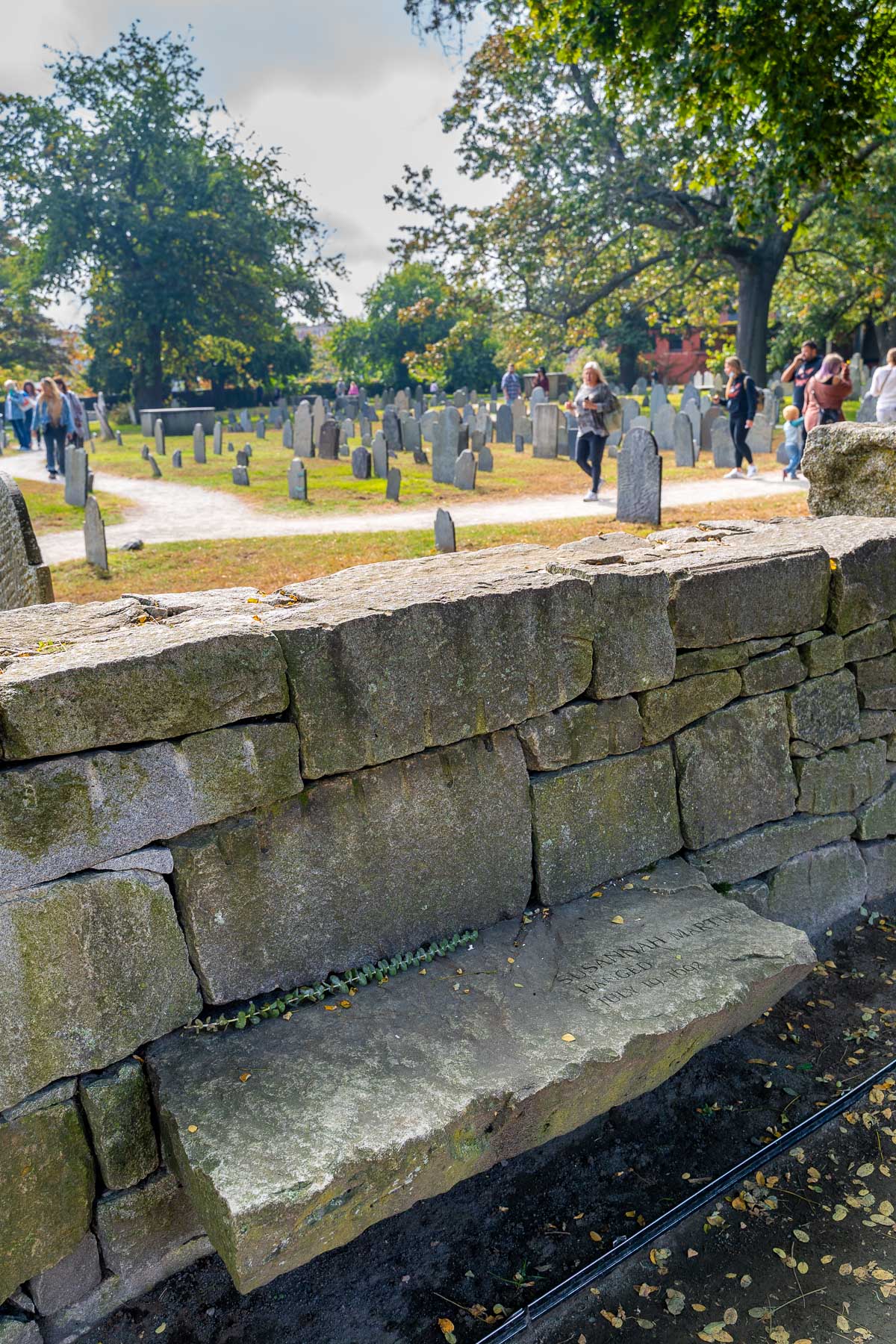 View of the Salem Witch Trials Memorial with a stone bench engraved with a victim’s name in the foreground and gravestones scattered across the grassy cemetery behind it.