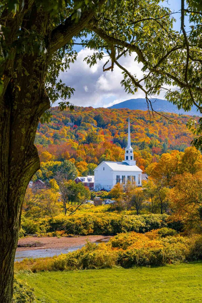 A white church with a tall steeple stands out against a backdrop of fall foliage in Stowe, Vermont, framed by tree branches in the foreground.