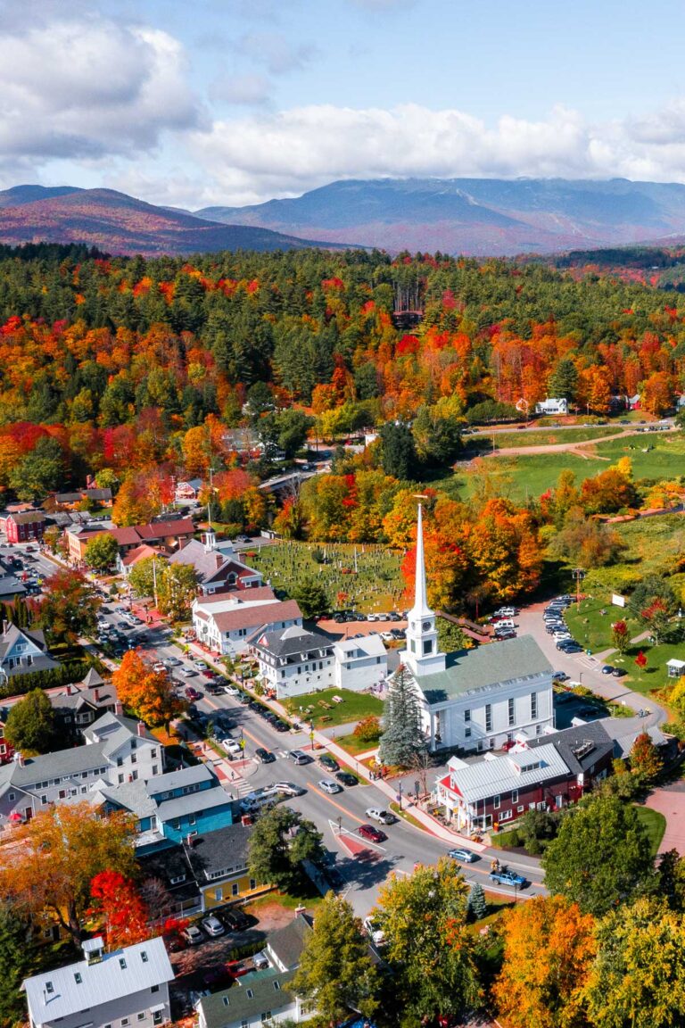 An aerial view of downtown Stowe, Vermont, showing colorful fall trees surrounding the white church and clustered houses below with mountain views in the distance.