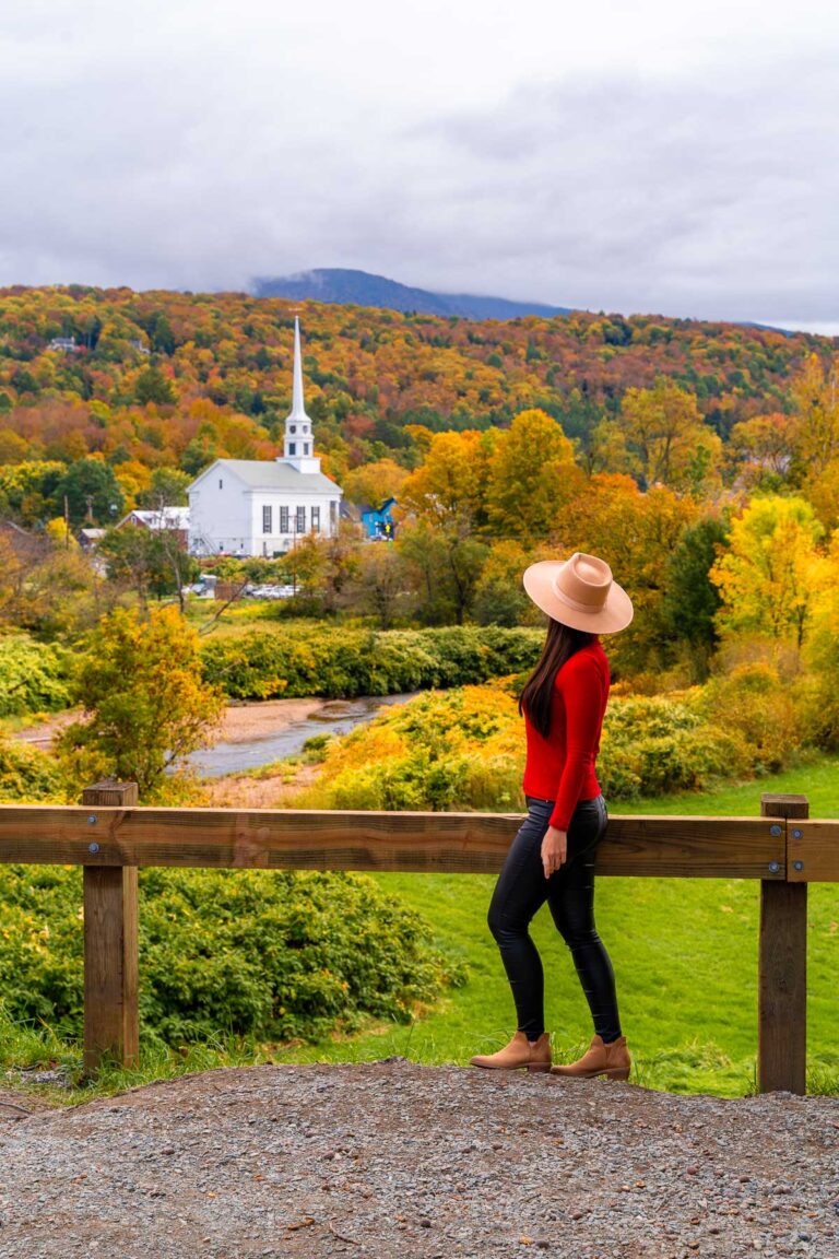 A woman in a hat and red sweater stands at a wooden railing with the white church in Stowe and colorful fall foliage on the hillside in the background.