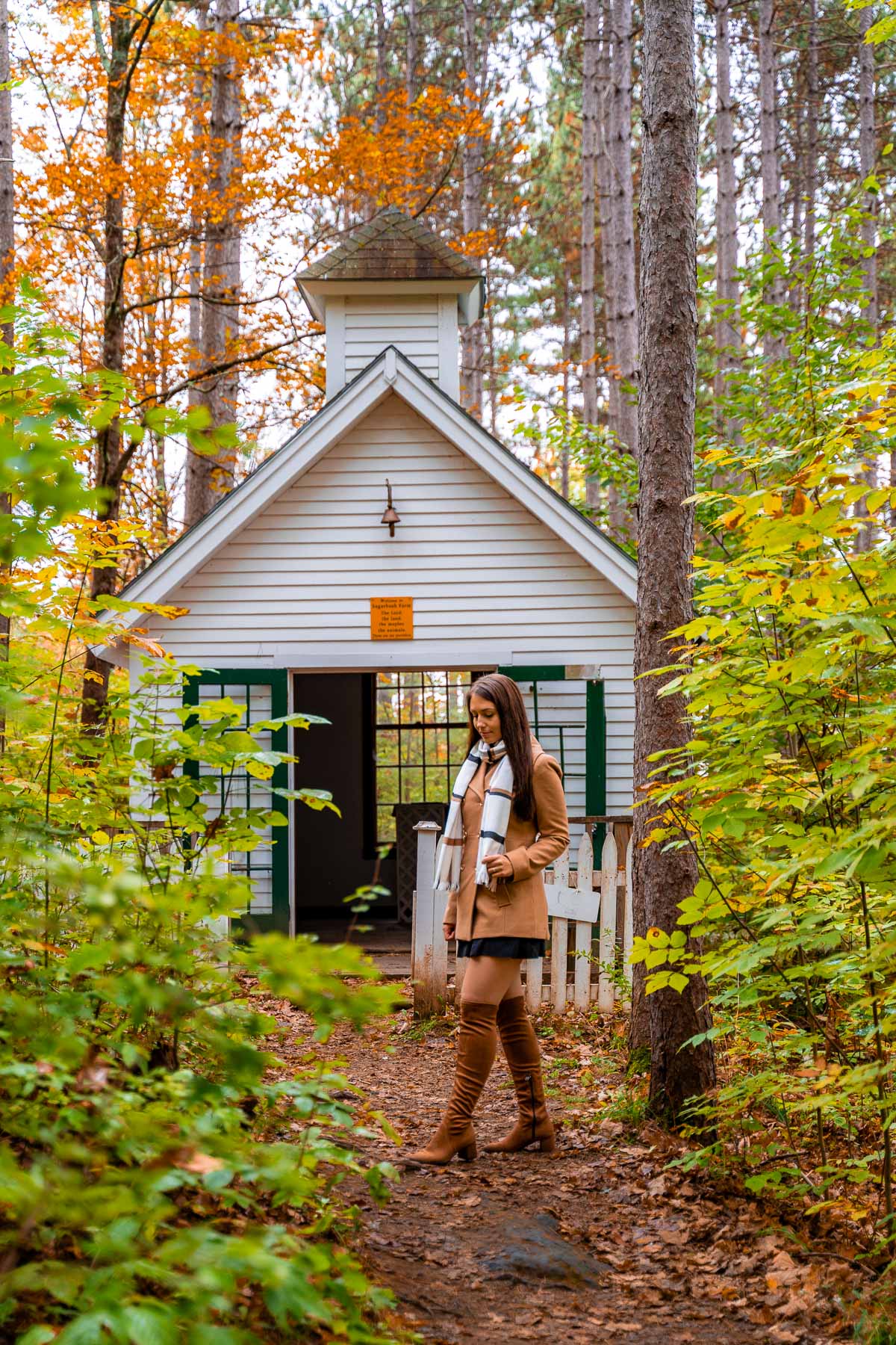 A small white chapel surrounded by fall foliage on the Maple Walk trail at Sugarbush Farm, with a woman walking in front of it.