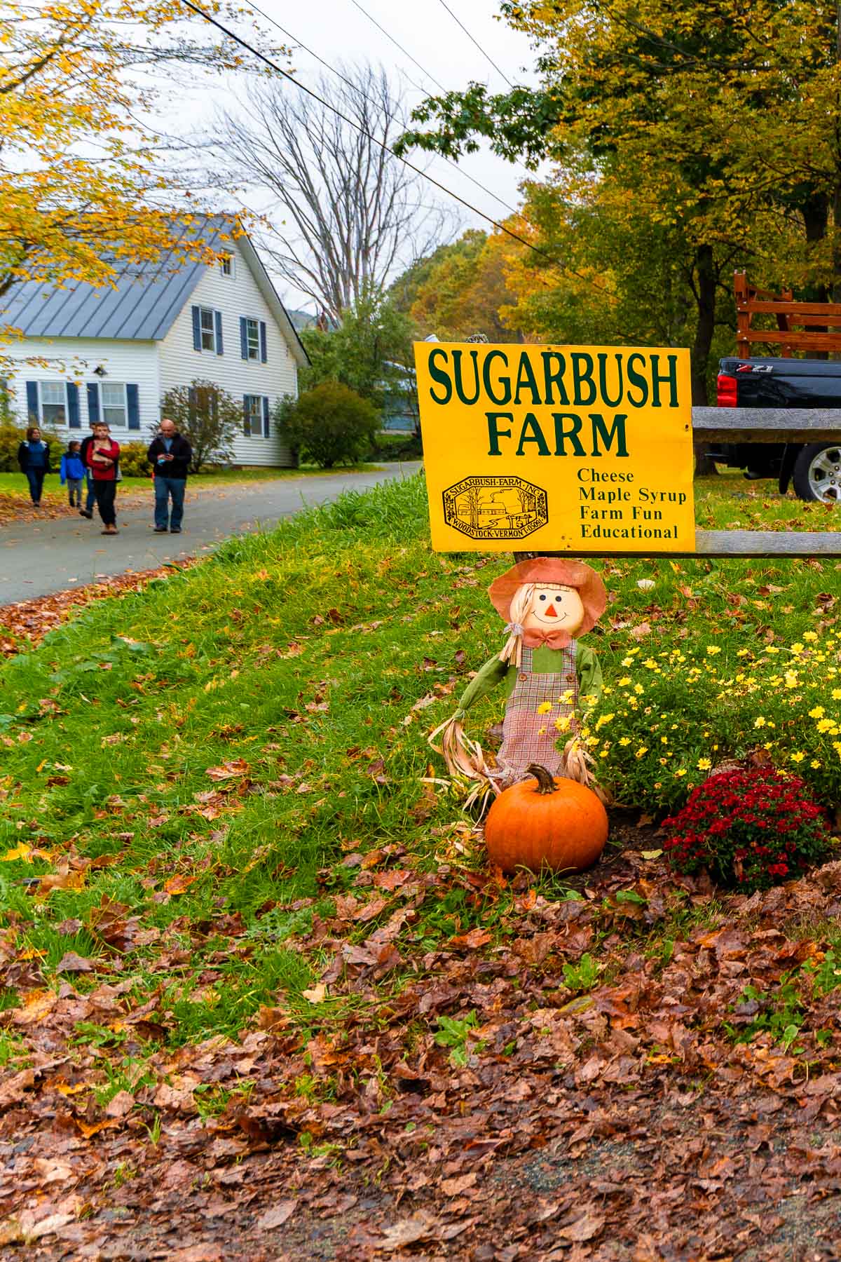 The entrance sign for Sugarbush Farm in Vermont sits on a grassy hill with a pumpkin and scarecrow decoration nearby and people walking along the road in the background.