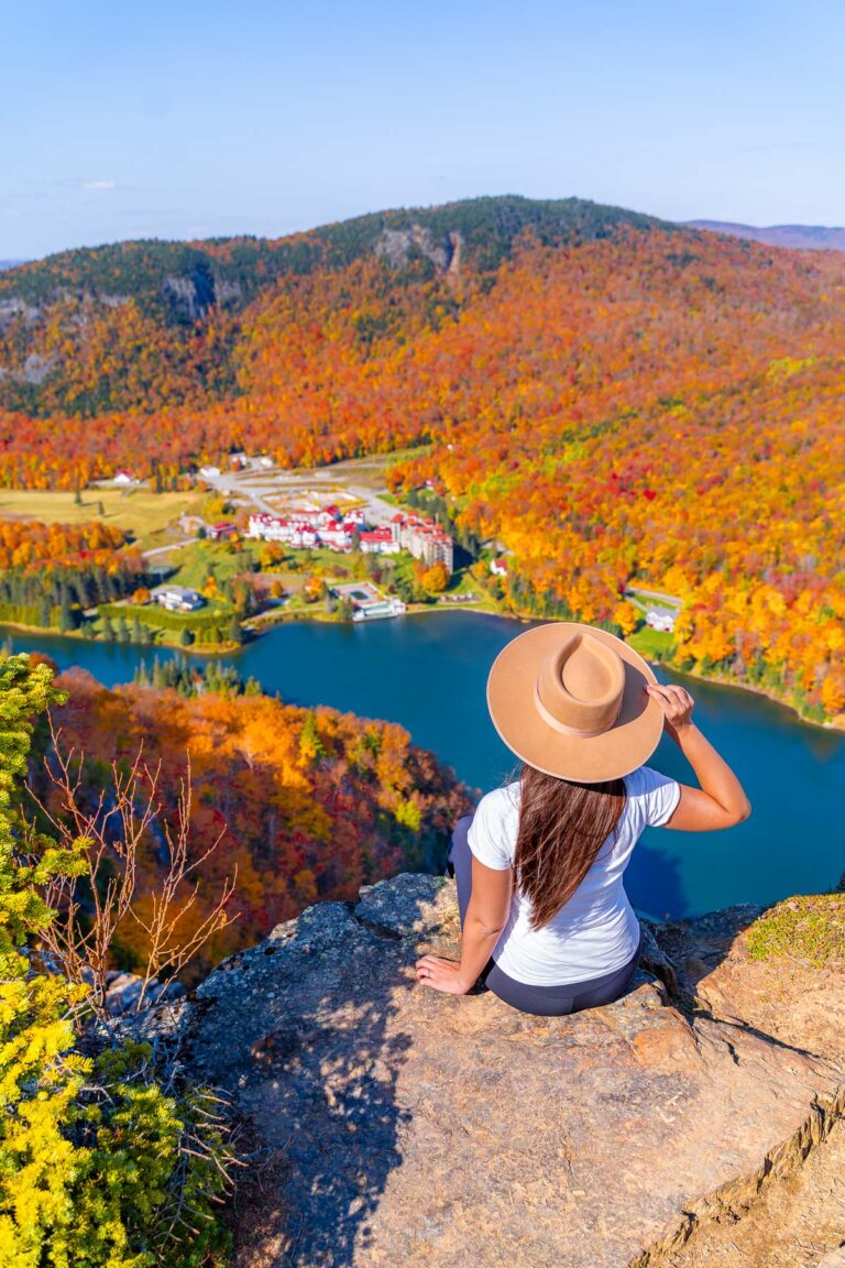 A woman sits on the edge of Table Rock overlooking a lake and vibrant fall foliage in Dixville Notch, New Hampshire with a view of The Balsams resort below.