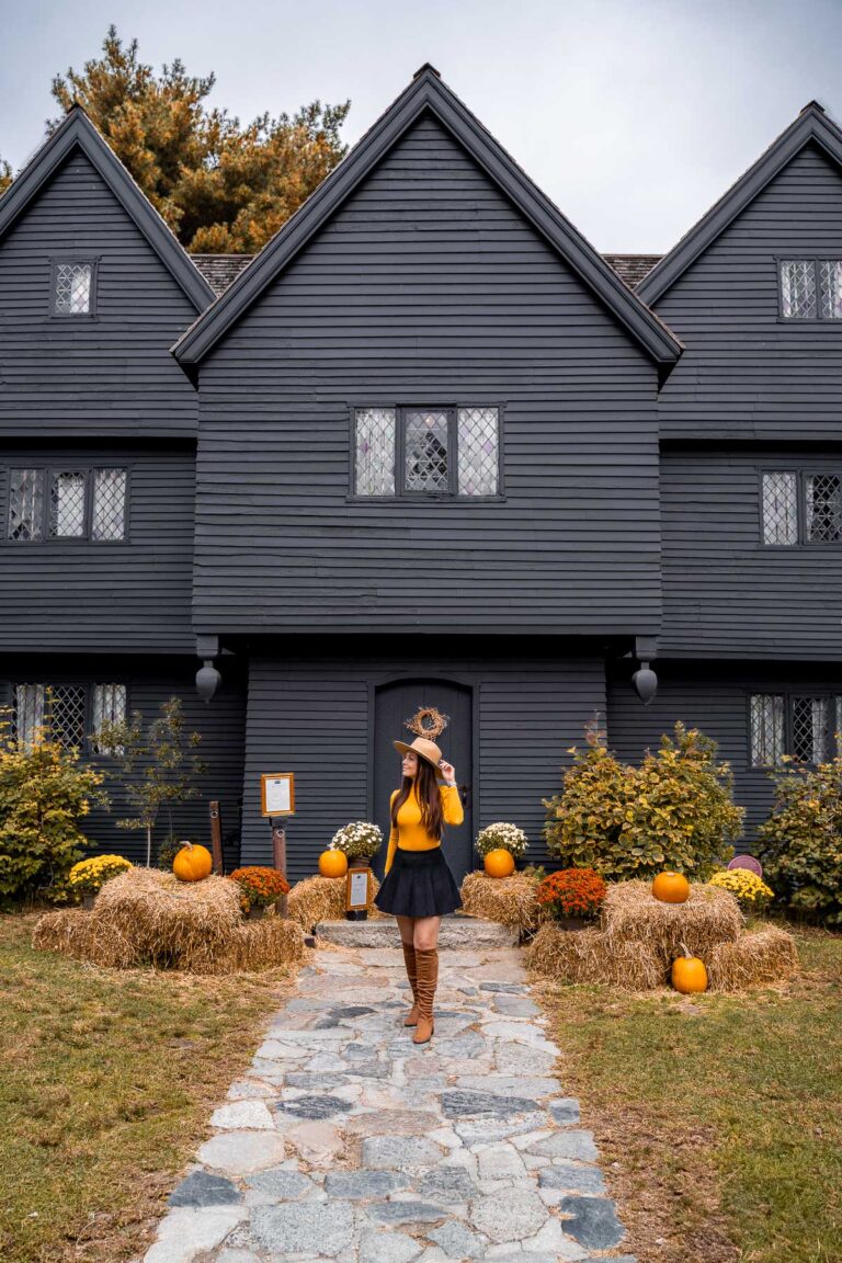 A woman in fall outfit stands on the stone path in front of The Witch House in Salem, surrounded by festive fall decorations.
