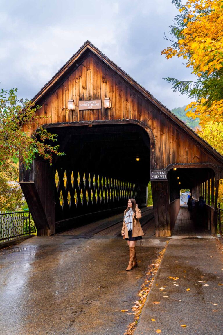 A woman walking in front of the rustic Woodstock Middle Covered Bridge in Vermont on a wet autumn day, with colorful fall trees in the background.