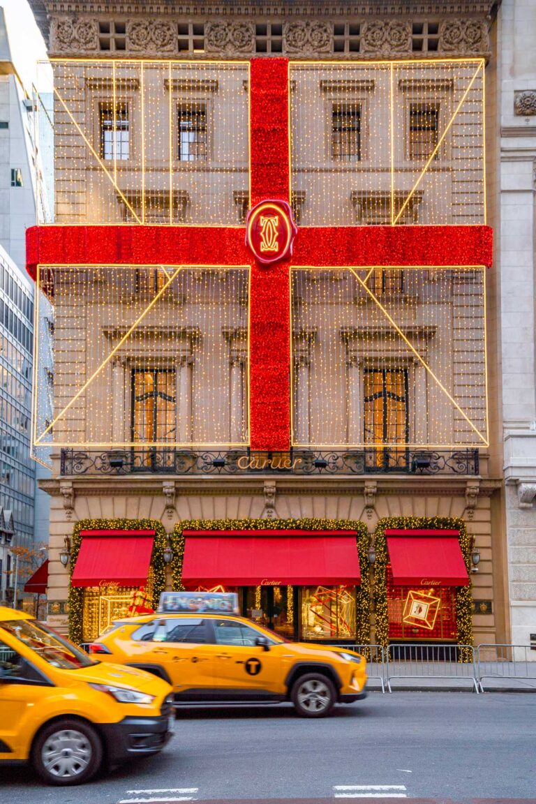 The Cartier Fifth Avenue building in New York decorated like a giant gift box with red ribbon-style lights and festive garlands, with yellow taxis passing in front.