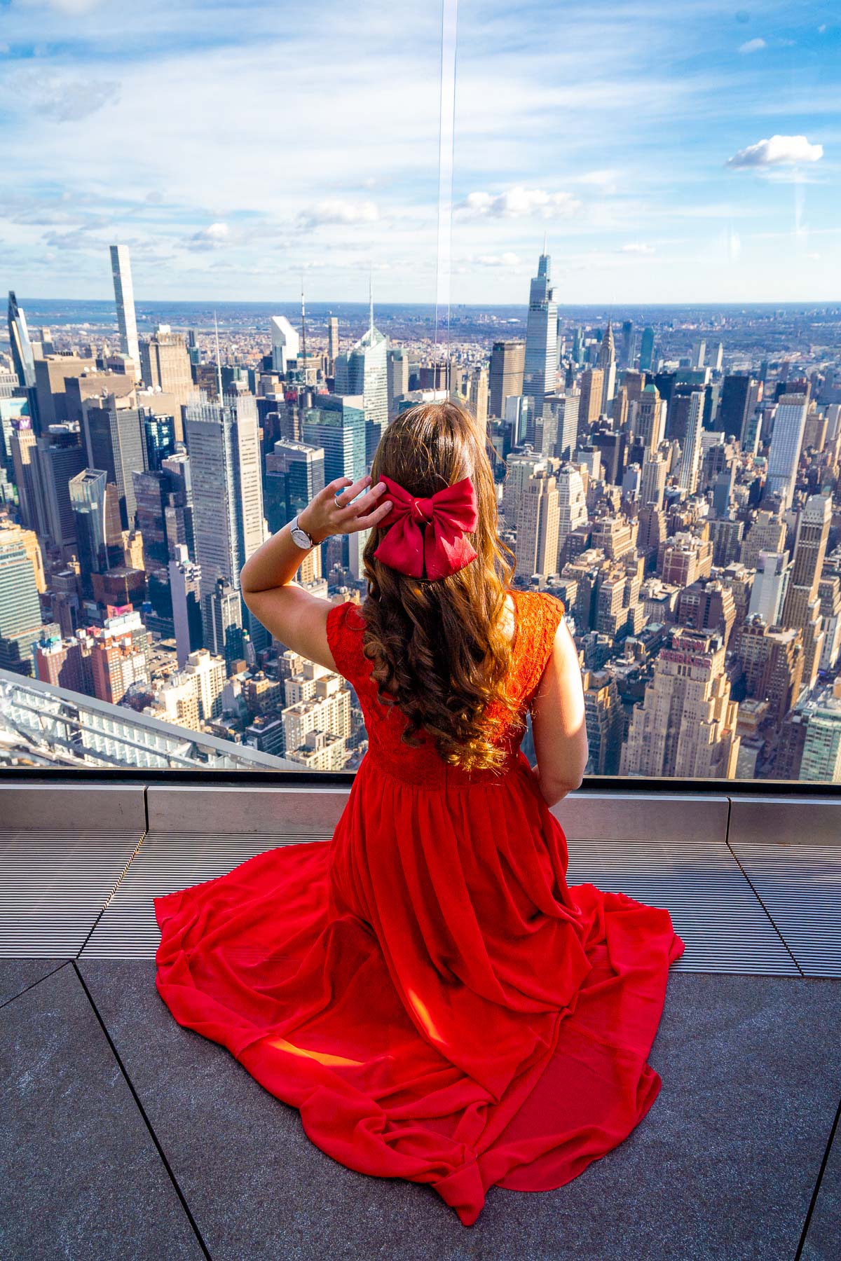 A woman in a red dress with a bow in her hair sitting at Edge in New York, looking out at the city skyline.